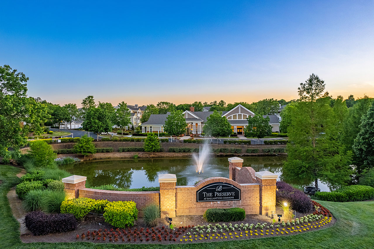 Scenic aerial view at sunset capturing an apartment complex, prominent monument sign, and a tranquil lake, bathed in warm, golden hues of the evening sky. Captured by Arch Angle Media